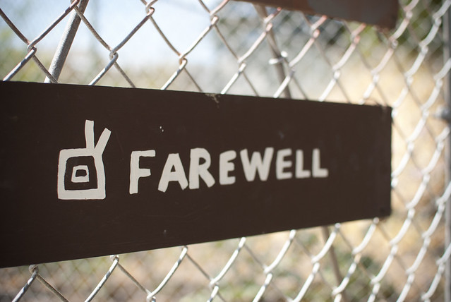 A sign reading ‘Farewell’. It’s white, hand-painted text on a dark brown/black background. The sign hangs on a chainlink fence.